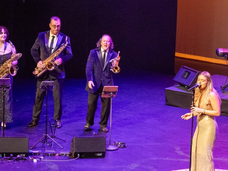 Image of A band performs on stage: a woman sings into a microphone at the front, with a saxophonist, a trumpeter, a second saxophonist, and a keyboard player playing behind her. All are dressed formally under bright stage lighting.