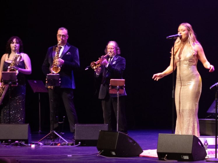 Image of A woman in a gold dress sings into a microphone on stage while three musicians beside her play saxophones and a trumpet. All are dressed formally, with stage lights highlighting the performers.