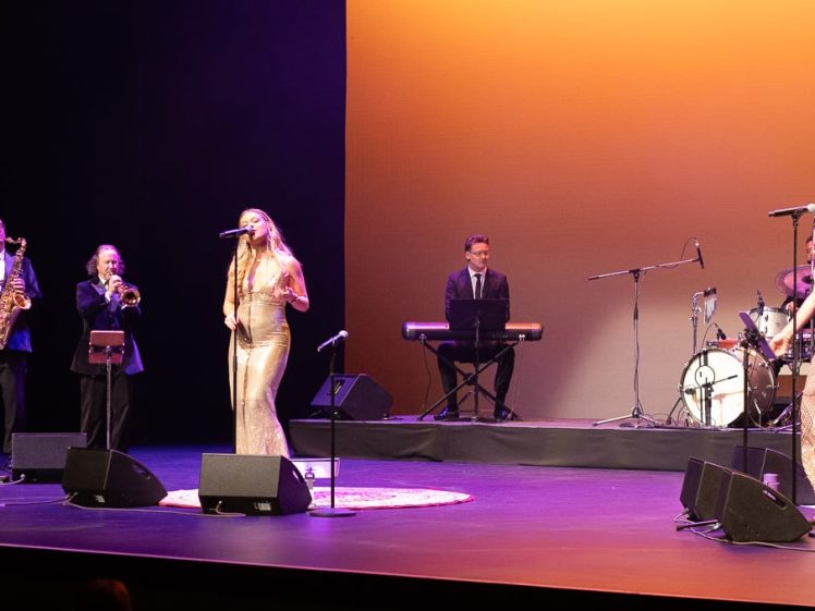 Image of A band performs on stage with singers in shimmering dresses and musicians playing saxophone, trumpet, keyboard, and drums against a warm, orange-lit backdrop.