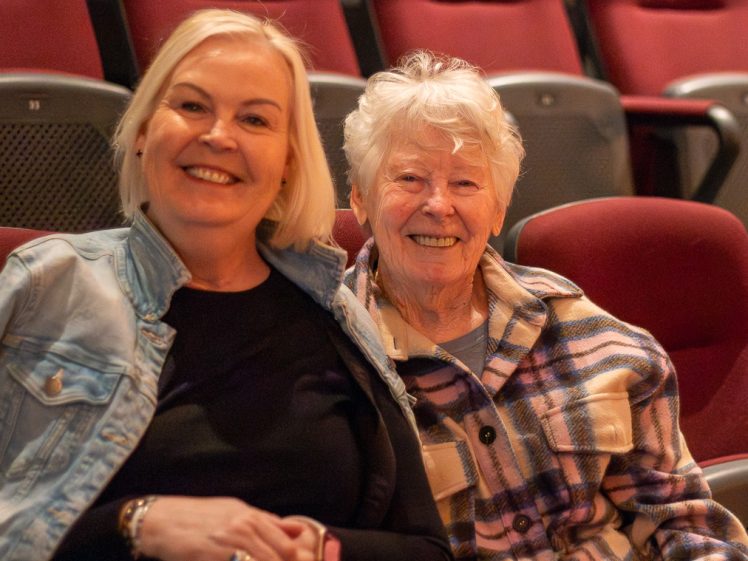Image of Two older women sit close together, smiling at the camera in a theatre with red seats. One wears a checked shirt; the other, a light denim jacket over a dark top. The background shows empty theatre seats.