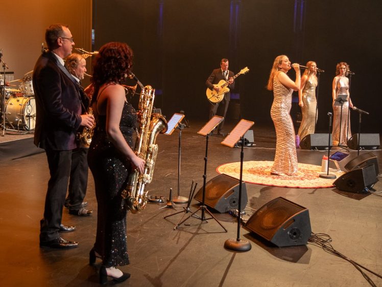 Image of A band performs on stage with singers in sparkly dresses and musicians playing saxophones, guitar, and drums. The stage is warmly lit, and several microphones and music stands are visible.