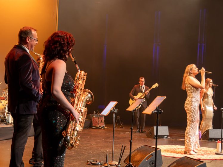 Image of A band performs on stage with saxophonists in the foreground, a drummer, guitarist, and three female vocalists in sparkling dresses singing into microphones, under warm stage lighting.
