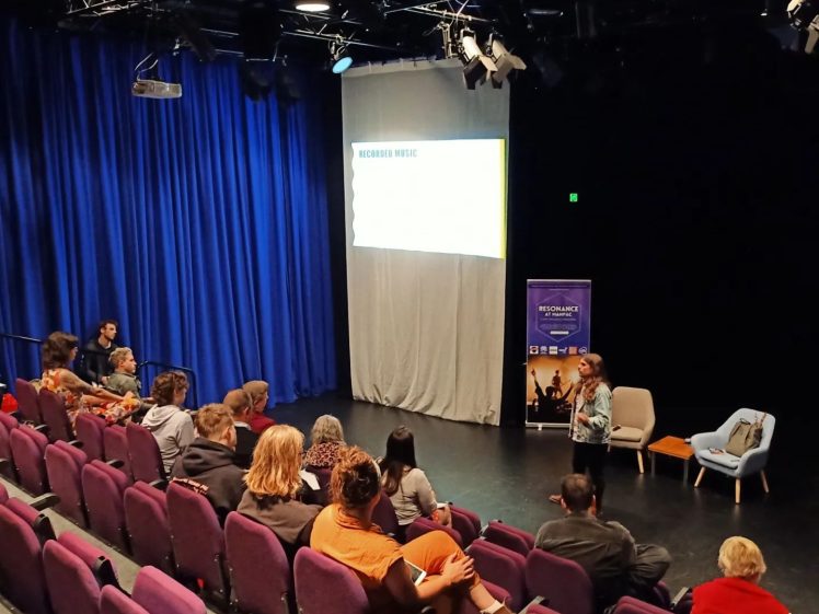 Image of A woman stands presenting in front of a small audience seated in purple chairs in a theatre. A projection screen displays information, and stage lighting highlights the speaker and blue curtains.