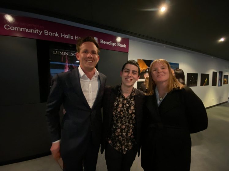Image of Three people stand smiling together indoors; one man in a suit, another in a floral shirt and blazer, and a woman in a black coat. They are posing under a sign for Bendigo Bank in a corridor with photos on the wall.