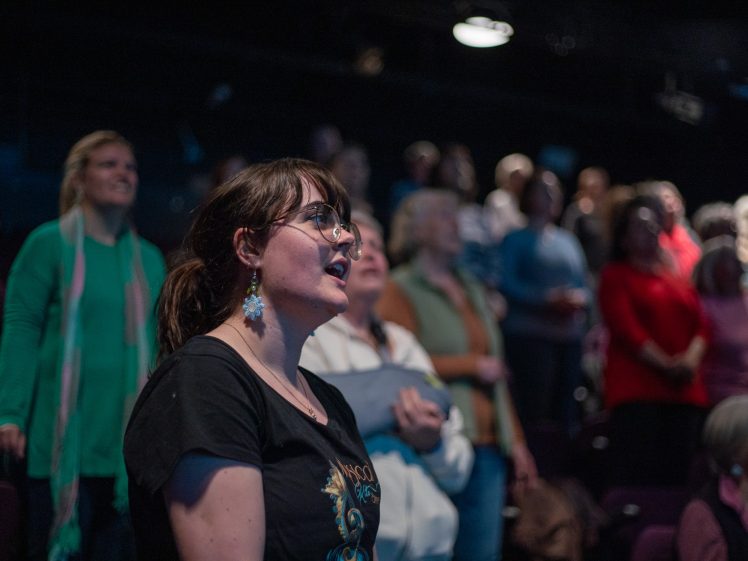 Image of A woman with glasses and earrings sits in focus among a group of people standing in a dimly lit theatre or auditorium, with blurred audience members in the background.