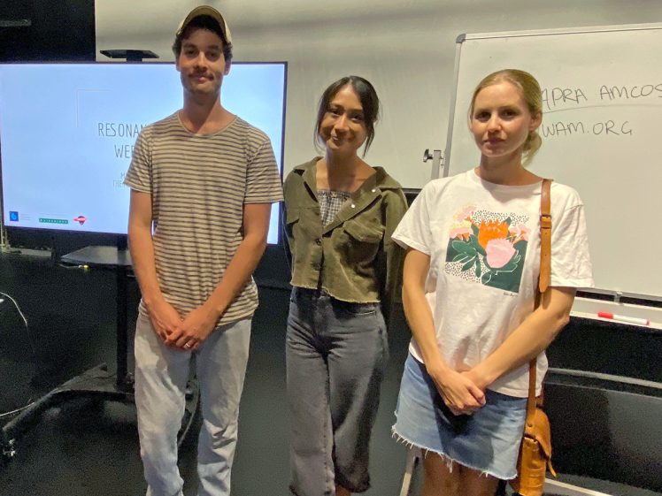 Image of Three people stand indoors, smiling at the camera. There is a large screen and a whiteboard behind them. The whiteboard has some writing on it, and the group appears to be in a classroom or meeting room setting.
