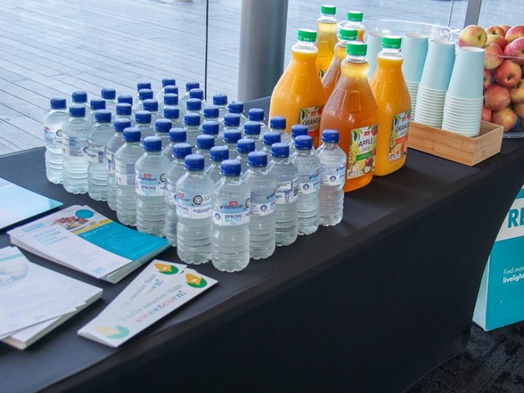 Image of A table with water bottles, juice bottles, paper cups, a container of apples, and brochures. A sign next to the table offers free apples and features healthy recipes.