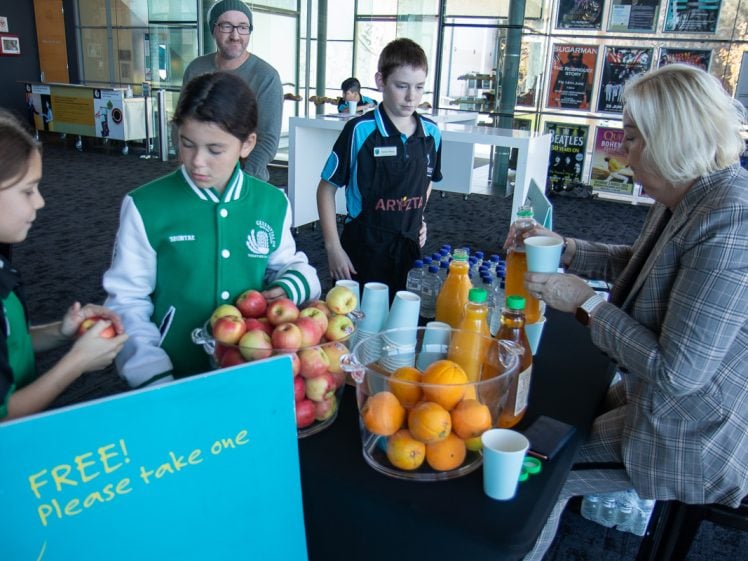 Image of Three children stand behind a table with apples, oranges, juice, and cups, offering refreshments to a seated woman. A sign reads FREE! Please take one. An adult watches from the background.