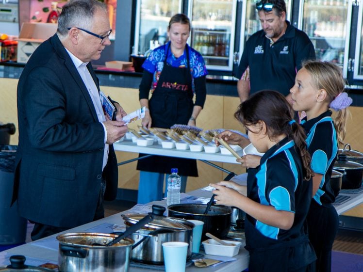 Image of A man in a suit talks to two young girls in matching uniforms at a table with food dishes. Other adults stand in the background at the indoor event. The girls hold notebooks and cutlery.