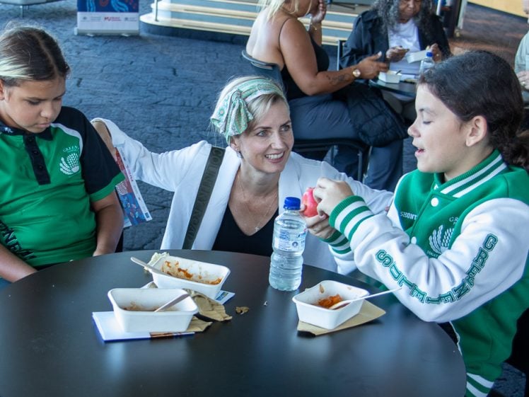 Image of A woman sits at a round table with two children, smiling and talking. They are eating a meal from rectangular bowls. The children wear green and black sports kits. Other people are seated in the background.