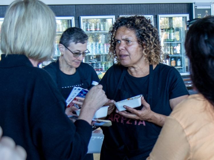 Image of Two women wearing aprons serve food in bowls to guests at an indoor event, standing in front of glass fridges stocked with drinks. One woman interacts with a guest holding a spoon.