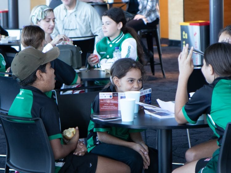 Image of A group of children in green sports kits sit at round tables in a canteen, eating and talking. Some adults are sitting in the background, and the atmosphere appears lively and sociable.