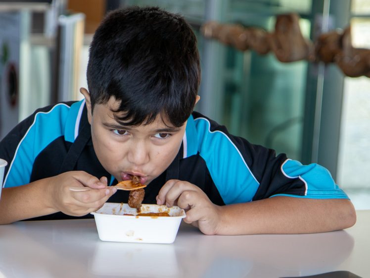 Image of A young boy with short dark hair and a blue and black shirt is sitting at a table, holding a fork and eating from a white takeaway container, looking down at his food with a focused expression.