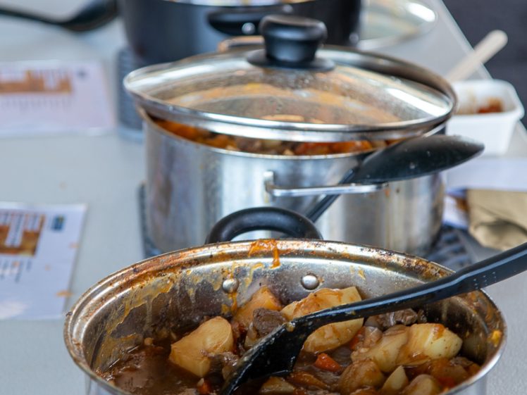 Image of Several pots of stew with ladles sit on a table, each containing a mixture of vegetables and meat, likely part of a cooking competition or tasting event. Flyers or papers are visible beneath each pot.