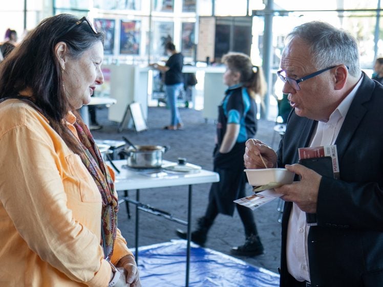 Image of A woman and a man converse indoors; the man wears glasses and holds a bowl with a spoon and papers, whilst the woman listens. Several people and tables appear in the background.