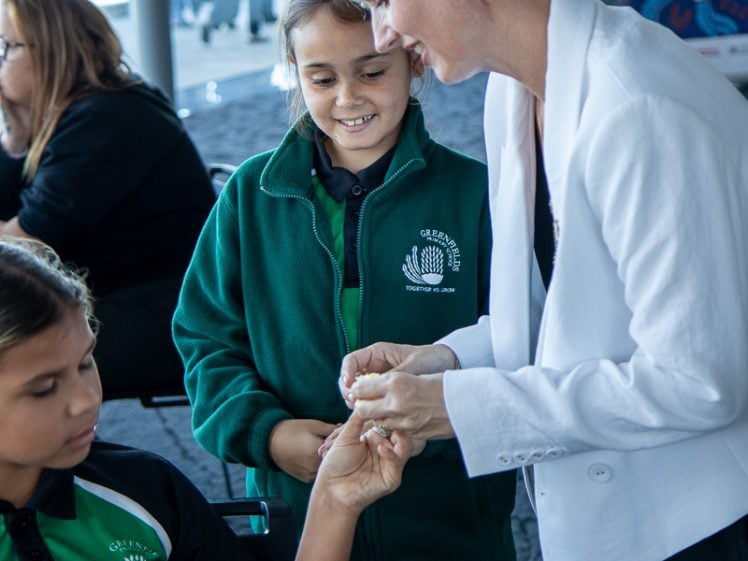 Image of A woman in a white jacket interacts with two children in green school uniforms, smiling and holding one child's hand. Other people sit in the background indoors by large windows.