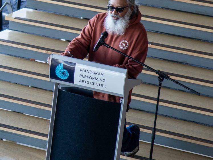 Image of A man with long grey hair and beard, wearing sunglasses and a brown hoodie, speaks at a podium labelled Mandurah Performing Arts Centre in front of wooden stairs.