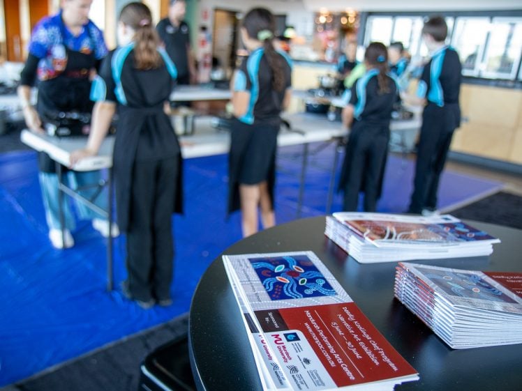 Image of Stacks of brochures about a sensory arts and science event sit on a table in the foreground, whilst children and adults participate in activities at tables in the blurred background.
