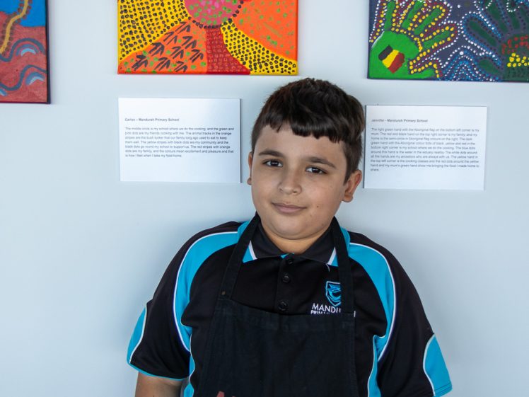 Image of A boy wearing a black apron with ARYZTA on it and a blue-and-black polo stands in front of a white wall displaying colourful abstract artworks and descriptions.