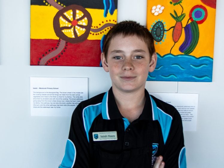 Image of A boy with short brown hair, wearing a black and teal school uniform with a name tag, stands with arms folded in front of colourful Aboriginal-style artwork displayed on a wall.