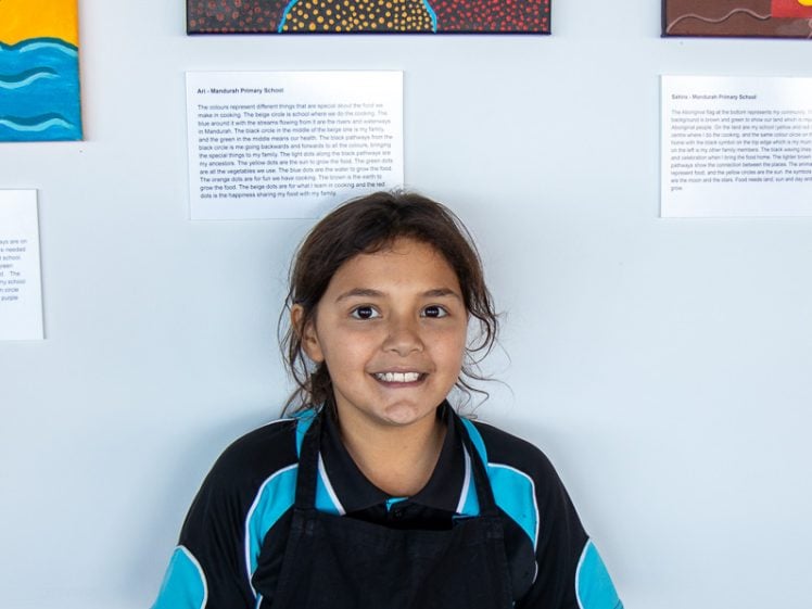 Image of A smiling child with dark hair, wearing a black apron with ARYZTA written on it, stands in front of colourful dot paintings displayed on a white wall.