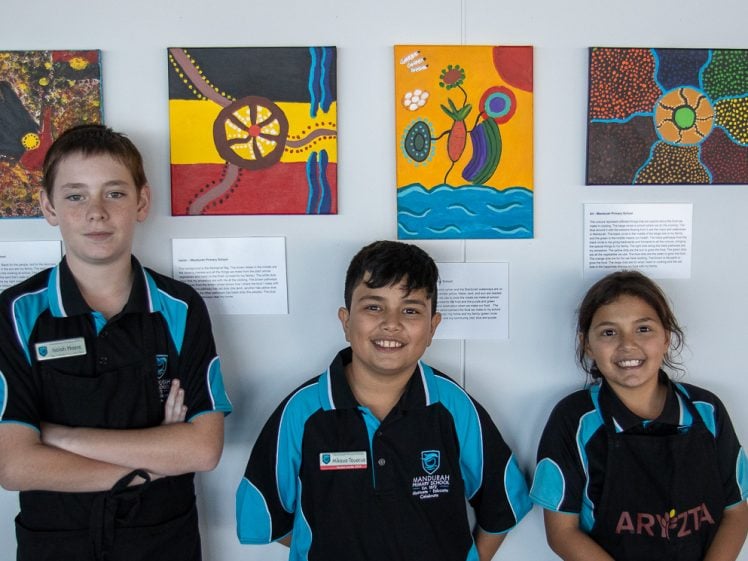 Image of Three children in school uniforms stand smiling in front of a wall displaying colourful Aboriginal-style paintings and informational plaques.