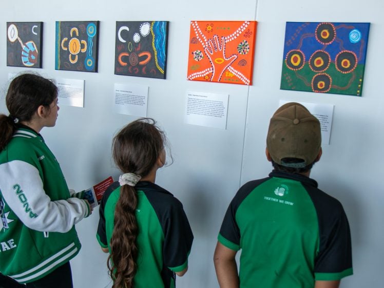 Image of Three children in green and black uniforms view colourful, abstract paintings and informational plaques displayed on a white wall in an art gallery.