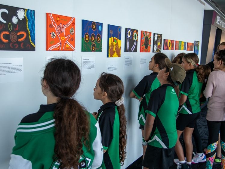 Image of A group of children in green uniforms view colourful artwork displayed on a white wall, with accompanying descriptions, in what appears to be a gallery or exhibition space.