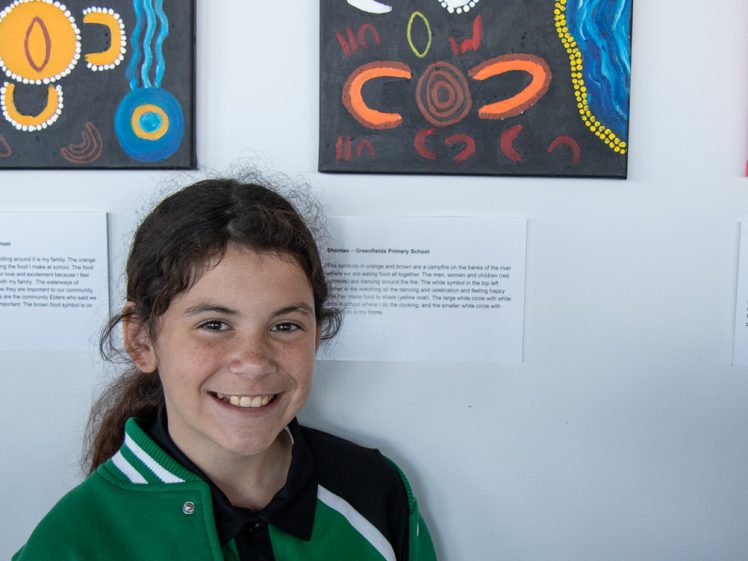 Image of A smiling girl with long brown hair, wearing a green and white jacket, stands in front of a wall displaying colourful Aboriginal-style artwork and descriptive labels.