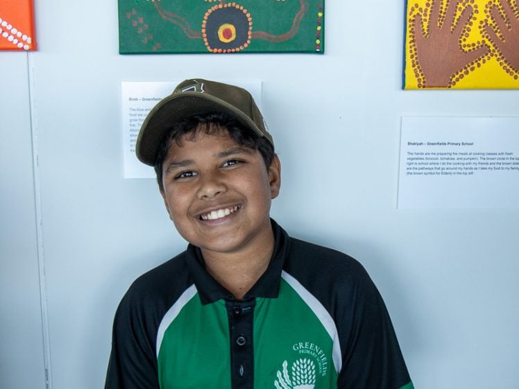 Image of A smiling boy wearing a green and black polo shirt and a brown cap stands in front of colourful Aboriginal-style artwork displayed on a white wall.