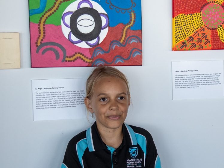 Image of A young girl in a black and teal Mandurah Primary School shirt stands smiling in front of a wall displaying colourful abstract paintings and information cards.