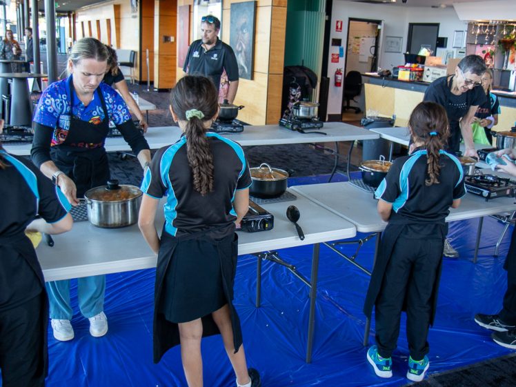 Image of Several children in matching uniforms stand around tables with cooking equipment, stirring pots, while two adults supervise inside a bright indoor space. The floor is covered with a blue tarpaulin.