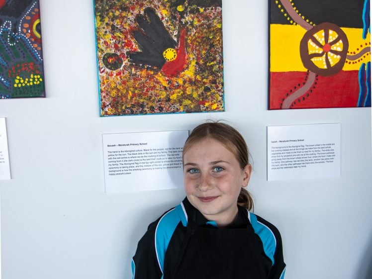 Image of A young girl in a black and blue shirt stands smiling in front of two colourful Aboriginal art paintings displayed on a white wall. She stands beneath an artwork and text label.