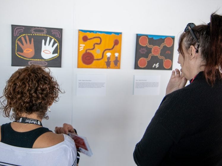 Image of Two people stand in front of a white wall, viewing colourful, abstract paintings and reading information cards at an art exhibition. Their backs are turned to the camera.