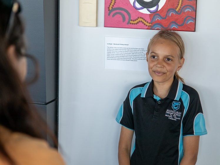 Image of A young girl in a Mandurah Catholic College uniform smiles while standing by artwork on a wall, speaking with another person whose back is to the camera. Art labels and colourful paintings are visible behind her.