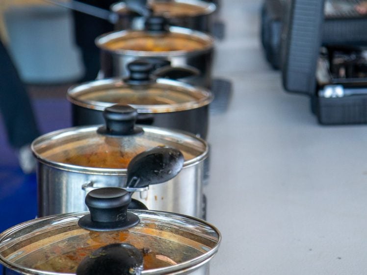 Image of A row of stainless steel saucepans with glass lids and black handles, filled with food, sits on a table. In the background, people are handling portable grills or waffle makers.
