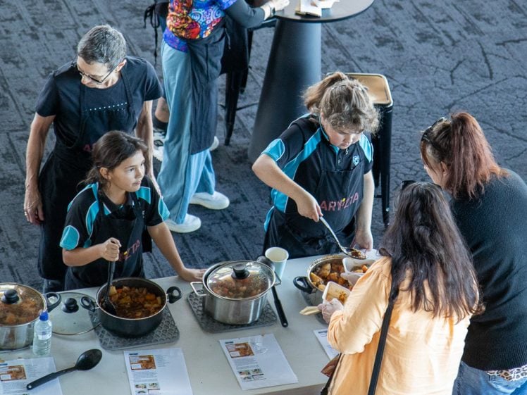 Image of Two children and two adults serve food from pots at a table with printed menus on it, while two other adults stand nearby. The scene appears to be a community or school event inside a large room.