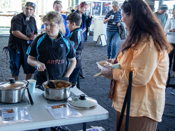 Image of A young person in a black apron serves food from a pot to a woman holding a bowl whilst a small crowd watches in a bright indoor setting. Other people are standing and observing in the background.