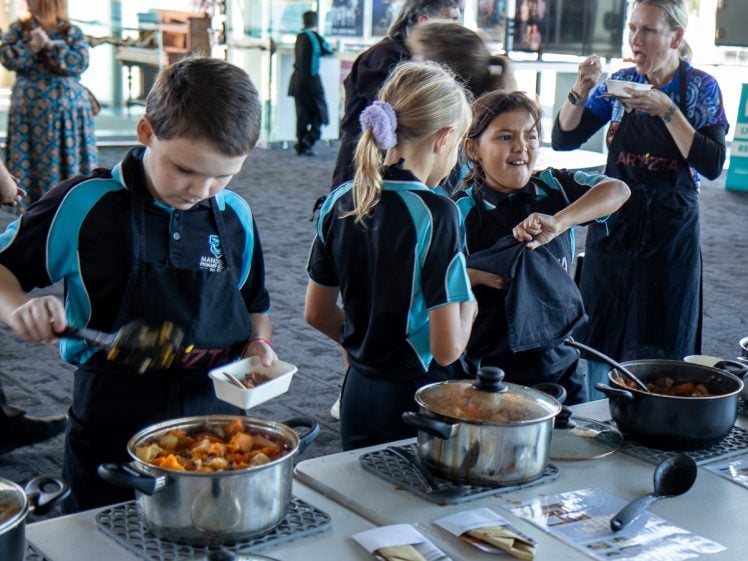 Image of Three children in matching uniforms serve food from pots into bowls at a table, whilst a woman in the background eats and others talk. The setting appears to be an indoor event or classroom.