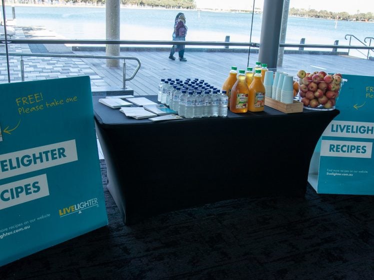 Image of A table with bottles of juice, water, and a basket of apples is set up indoors by large windows overlooking a waterfront. Two signs promote free LiveLighter recipes and encourage people to take one.