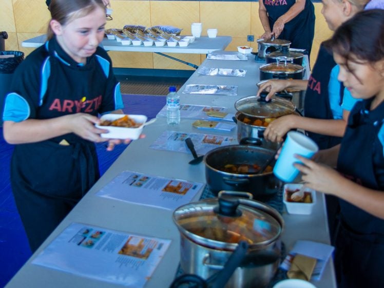 Image of Children and adults serve food from saucepans into bowls at a long table indoors, wearing aprons. Various cooking utensils and recipe sheets are visible on the table, with a kitchen and plates in the background.