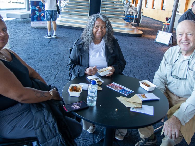 Image of Three people sit around a small round table, smiling at the camera. They have food containers, a water bottle, and papers on the table. A staircase and a few other people are visible in the background.
