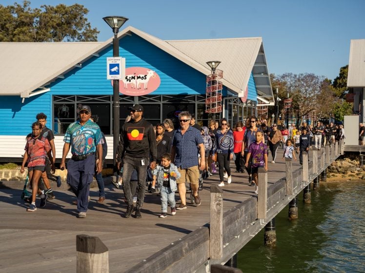 Image of A large, diverse group of people, including adults and children, walk along a wooden boardwalk next to blue buildings beside the water on a sunny day. Some wear colourful clothing with Aboriginal symbols.