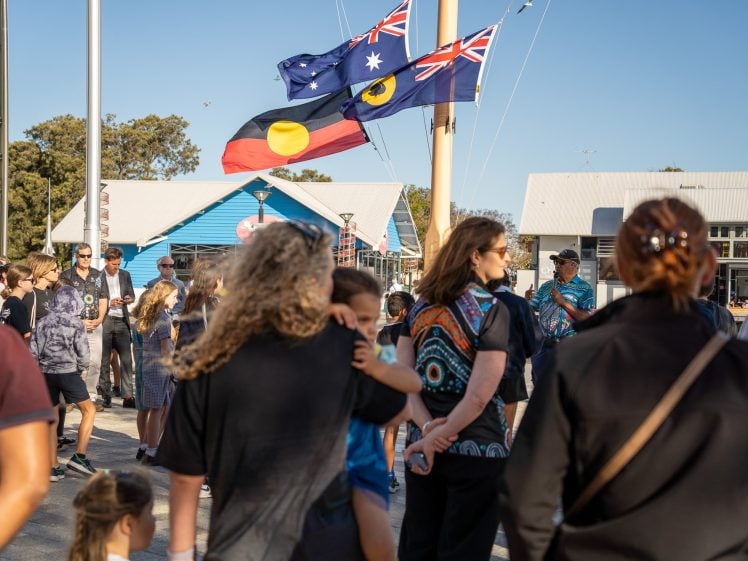 Image of A crowd gathers outdoors near flagpoles displaying the Australian, Aboriginal, and Torres Strait Islander flags. People of various ages stand and talk, with buildings and trees in the background under a clear sky.