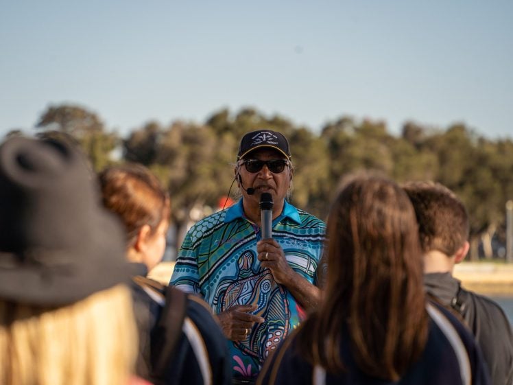 Image of A man wearing sunglasses, a cap, and a colourful shirt speaks into a microphone outdoors, facing a group of people with blurred faces. Trees and a body of water are visible in the background.