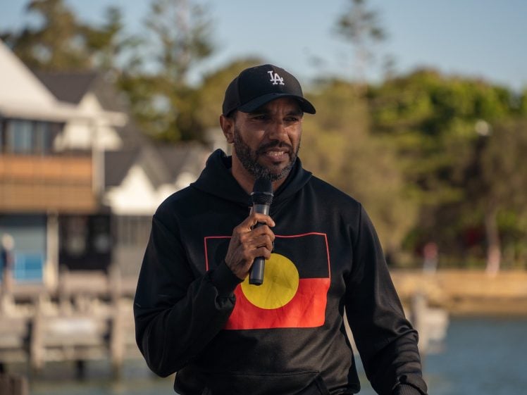 Image of A man wearing a black LA cap and a hoodie with the Australian Aboriginal flag design speaks into a microphone outdoors, with buildings, trees, and water in the background.