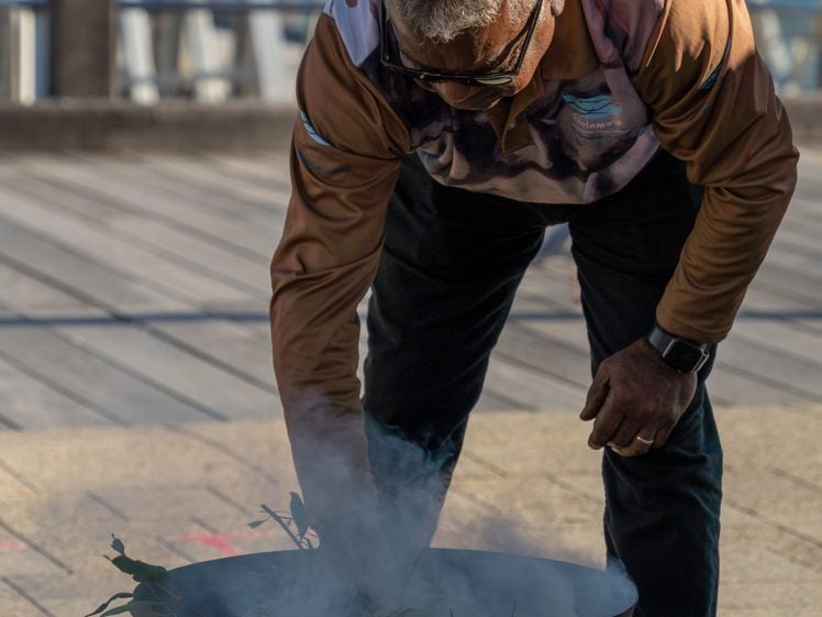Image of A man wearing sunglasses and a patterned shirt bends over a smoking fire pit with eucalyptus leaves, performing a traditional ceremony outdoors near a waterside.