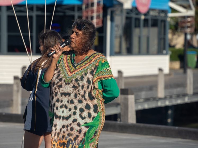 Image of An older woman in a colourful patterned dress speaks into a microphone outdoors. She stands near a young girl, with buildings and a waterfront in the background on a sunny day.