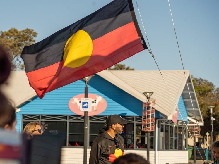 Image of A man wearing a black outfit with a yellow circle stands under an Aboriginal flag, which is waving in front of a blue and white building on a sunny day. Other people are partially visible.