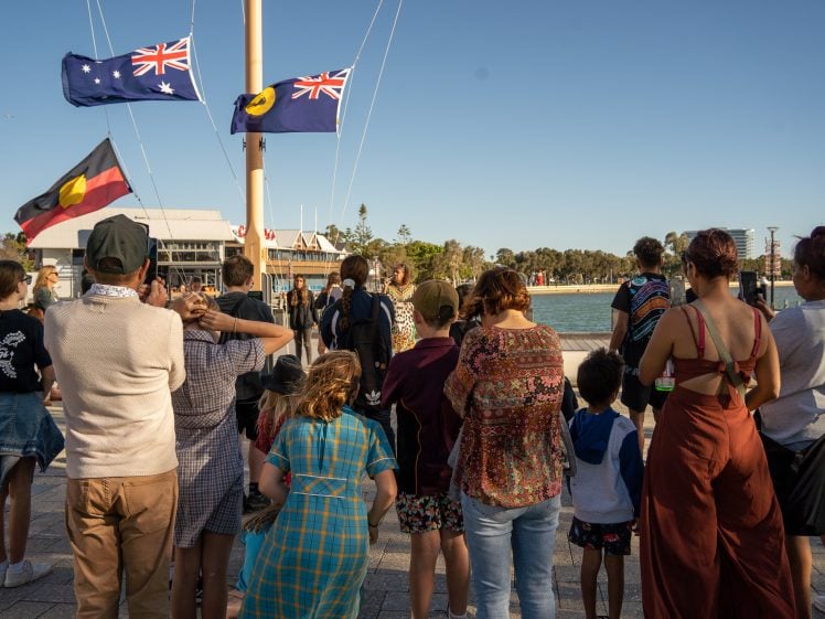 Image of A diverse group of people, including adults and children, stand together outdoors facing three raised flags near a waterfront under a clear sky. Some people hold or wear items with Aboriginal flag colours.